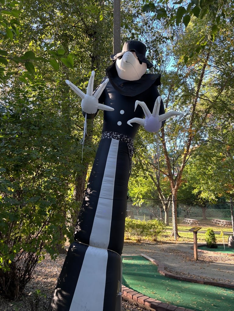 Tall cartoon character figure with black clothing and large white gloved hands standing next to mini golf course under trees
