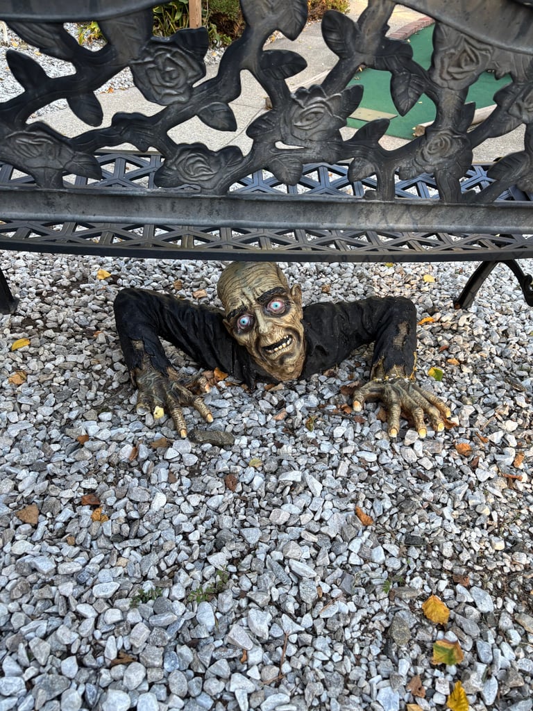 Bronze zombie statue with outstretched arms crawling under decorative metal fence on gravel ground