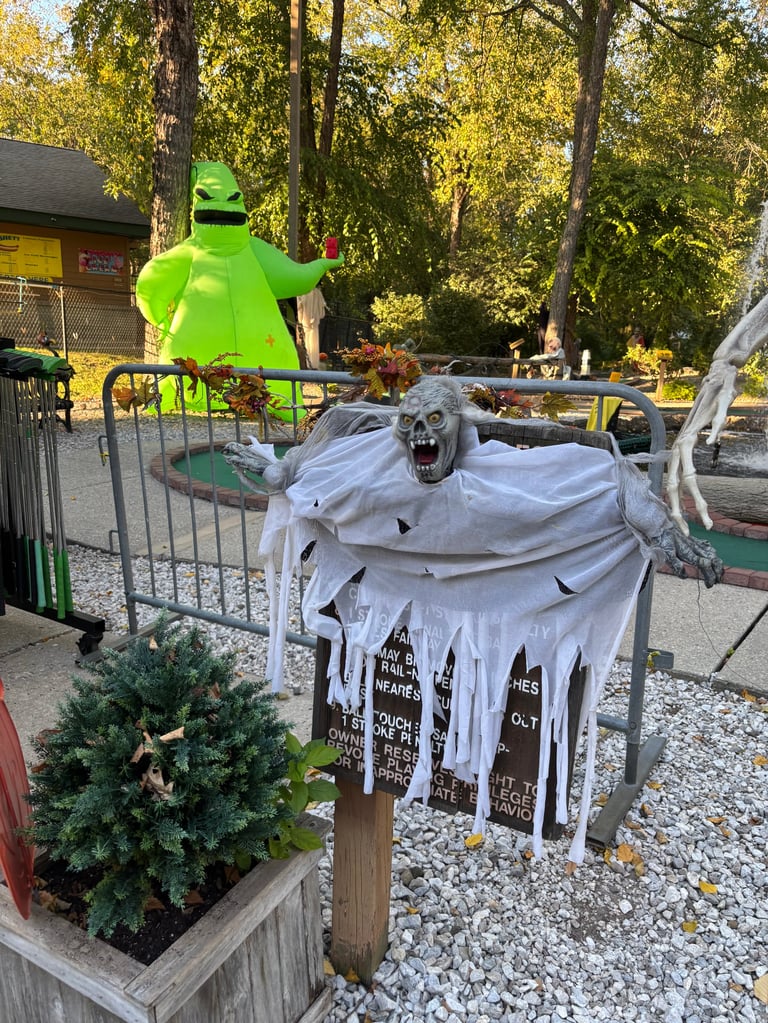 Outdoor miniature golf course with Halloween decorations including a ghostly figure in white tattered fabric and a person in bright green costume