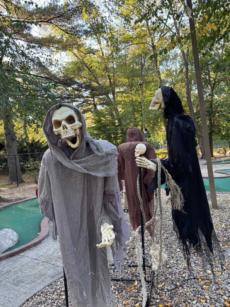 Three skeleton and grim reaper decorations at a mini golf course surrounded by fall foliage