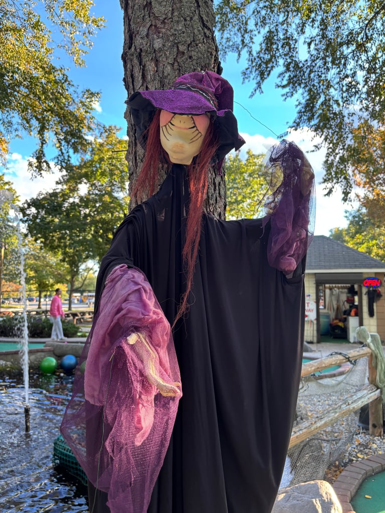 Witch decoration with black cloak, purple hat and long burgundy hair displayed on a tree trunk at an outdoor mini-golf course during autumn