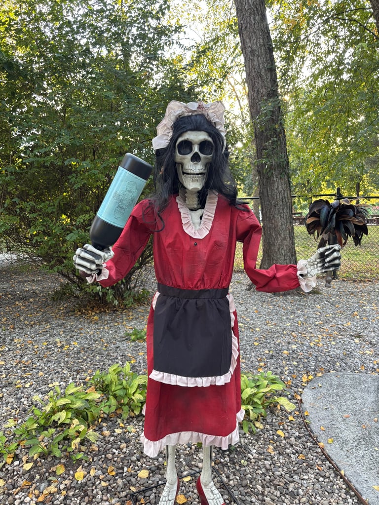 Skeleton figure in red dress holding a wine bottle, posed on a garden path with trees in background