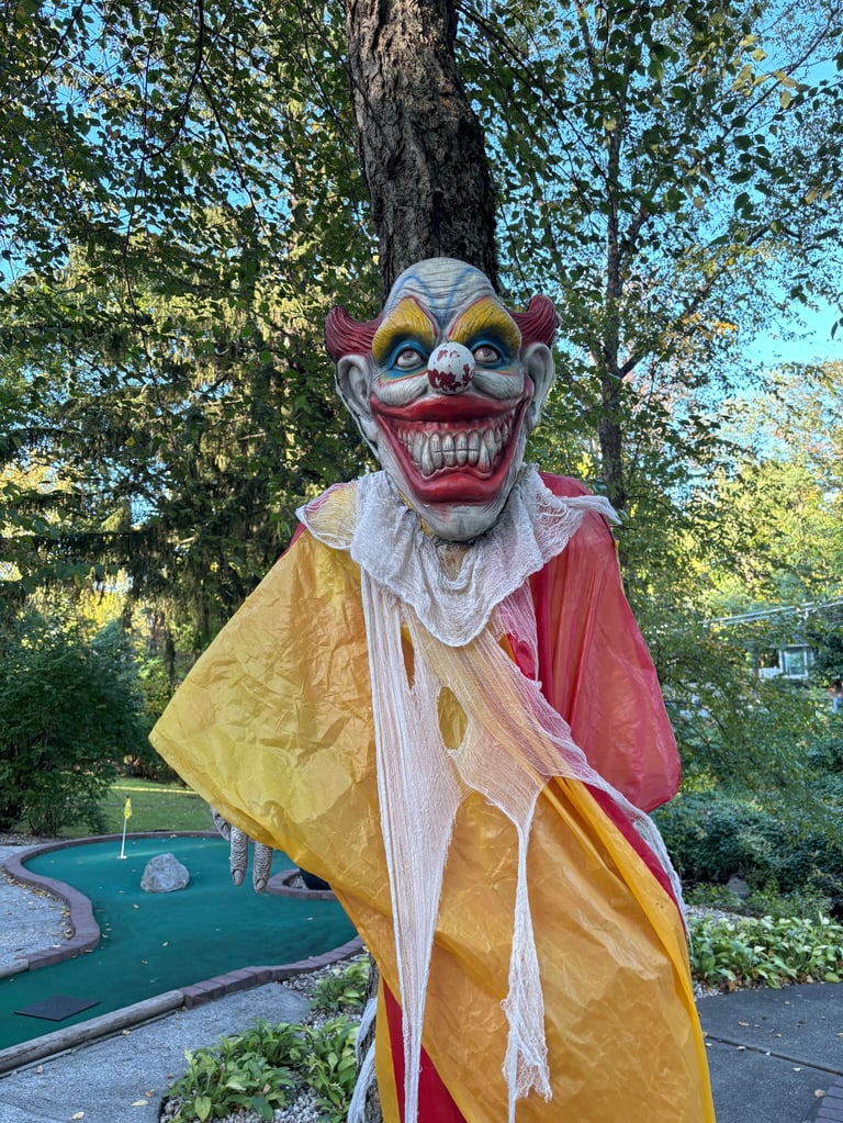 Creepy clown figure with exaggerated grin wearing yellow and red costume in a residential yard with mini golf course