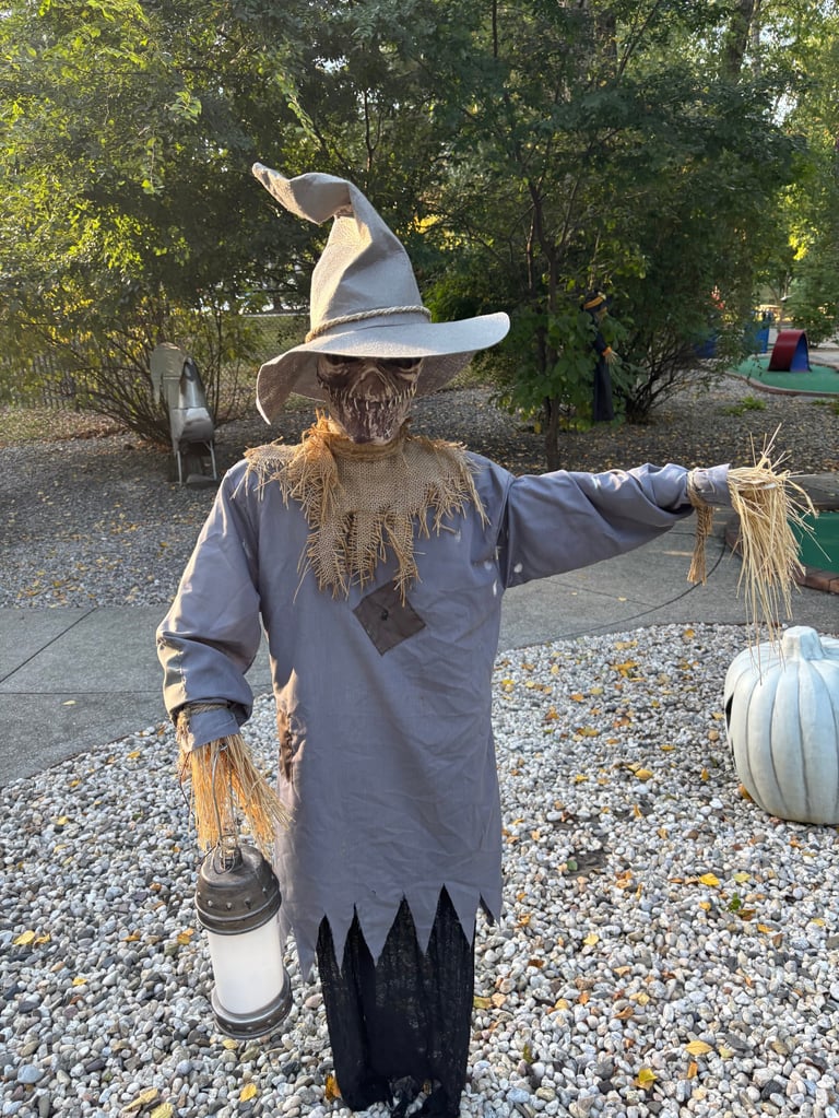 Scarecrow in witch hat holding straw and lantern in autumn garden display with pumpkin and trees