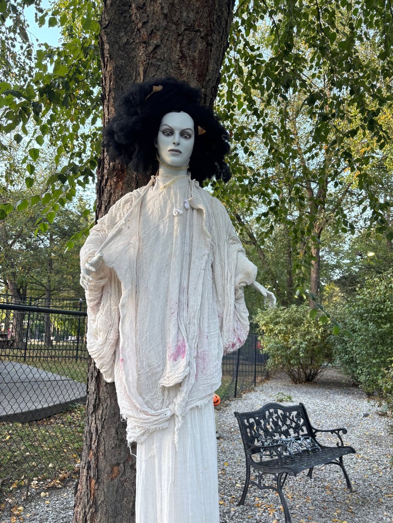 Ghostly figure with pale skin and dark hair wearing white draped fabric, leaning against a large tree trunk in an outdoor park setting